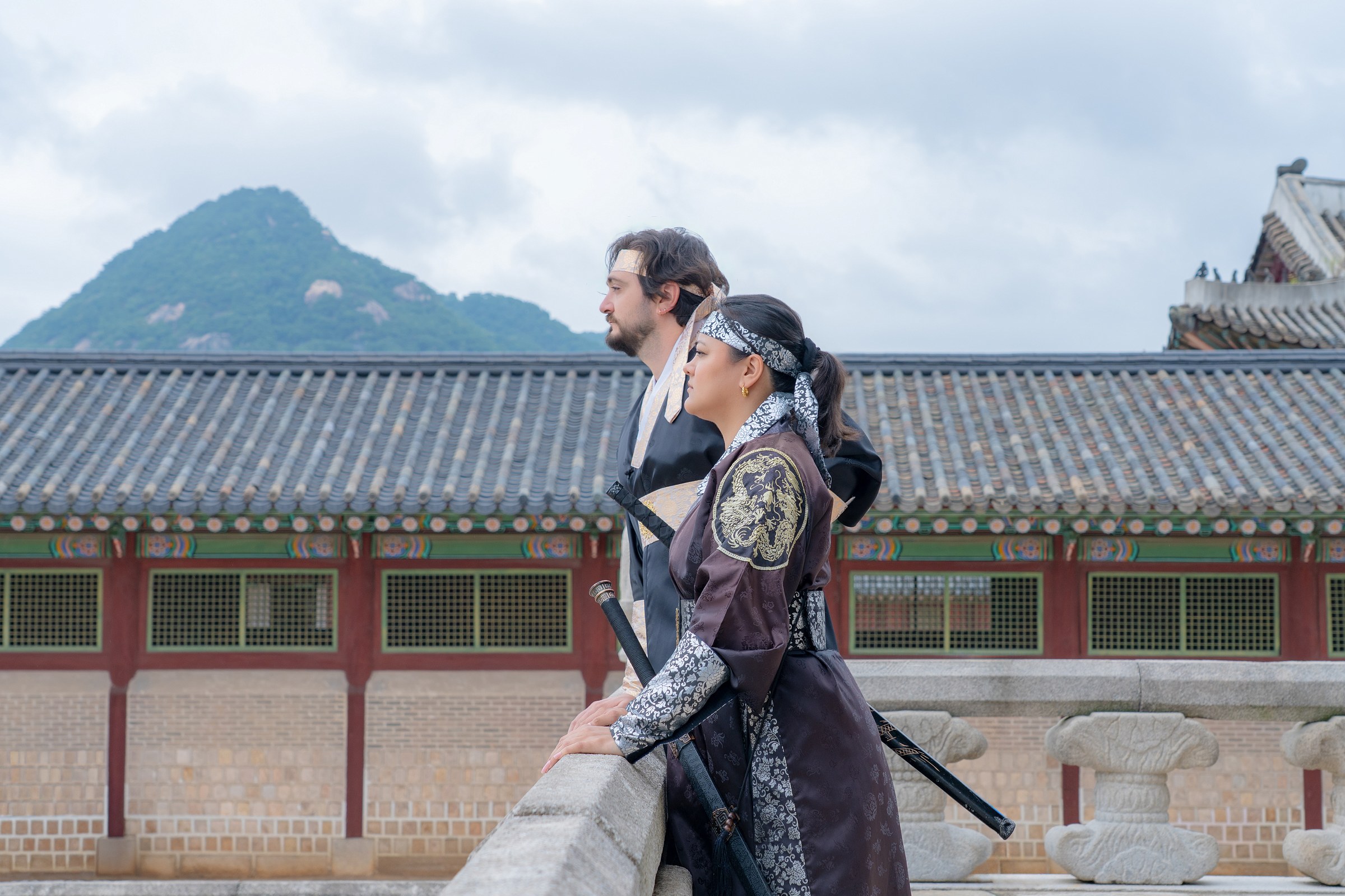 A couple in black hanbok walking beside a palace pavilion and mountain view at Gyeongbokgung