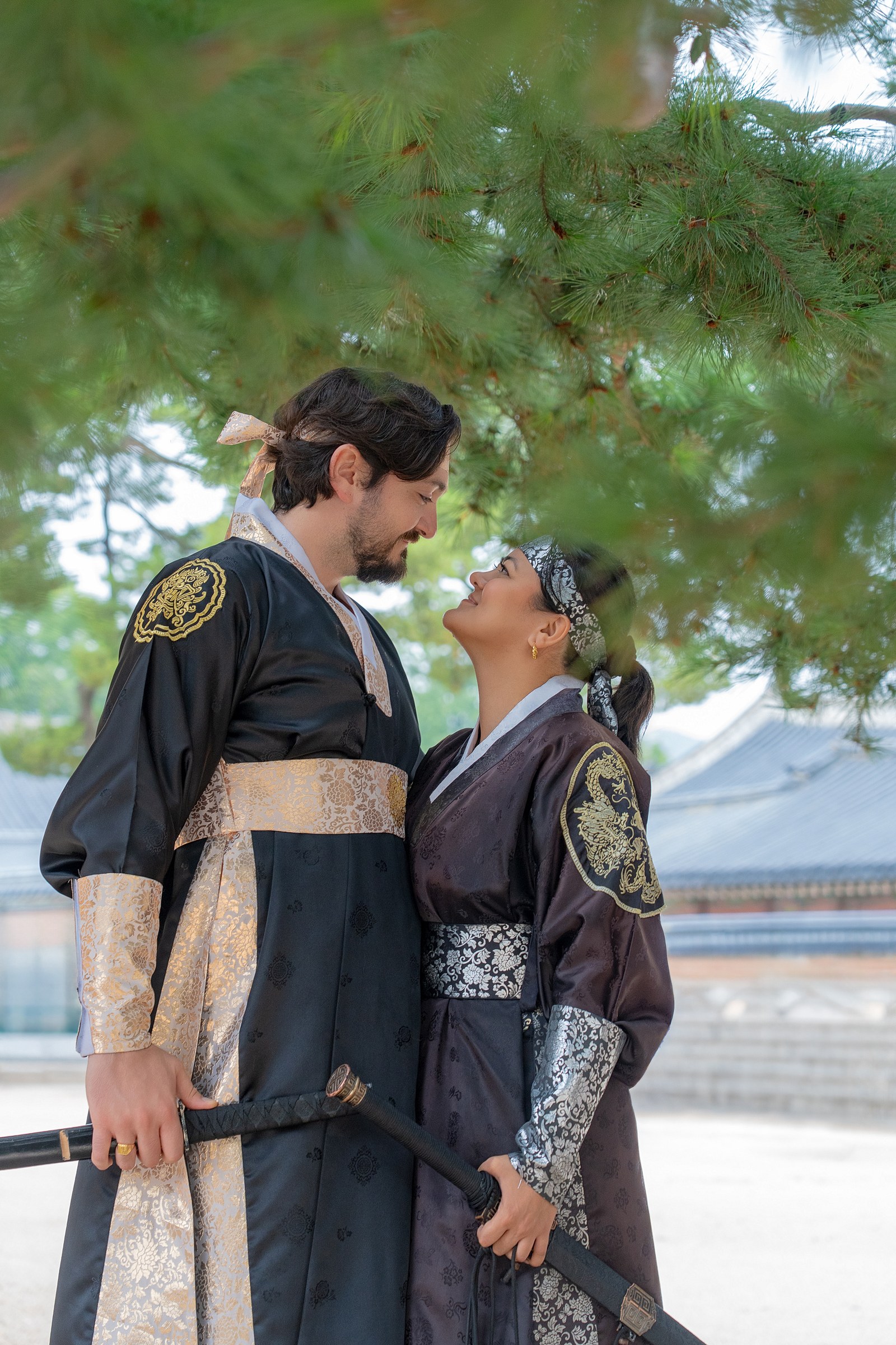 A close outdoor portrait of a couple in black hanbok beneath pine trees