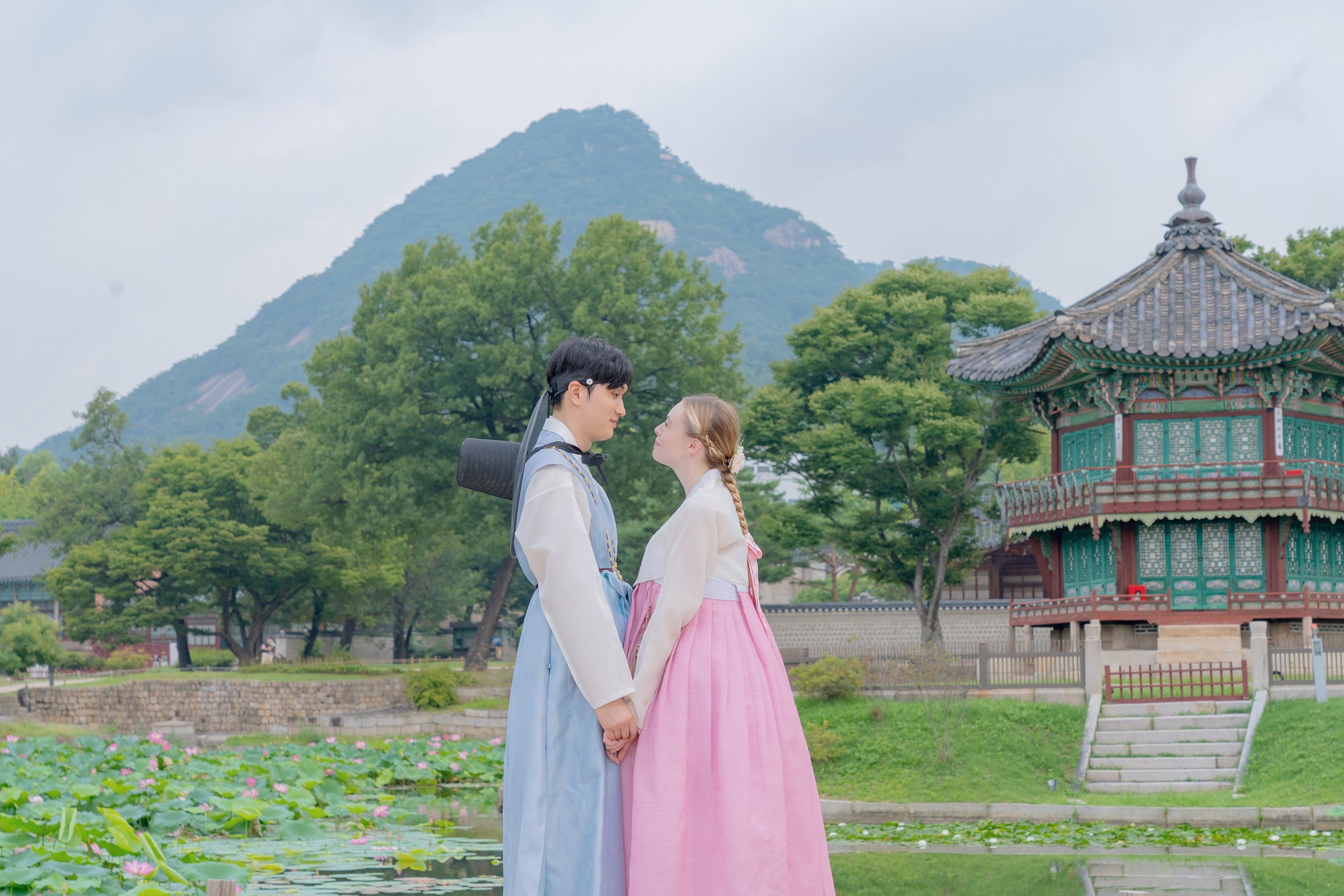 A couple in pastel hanbok holding hands beside a pond and pavilion at Gyeongbokgung
