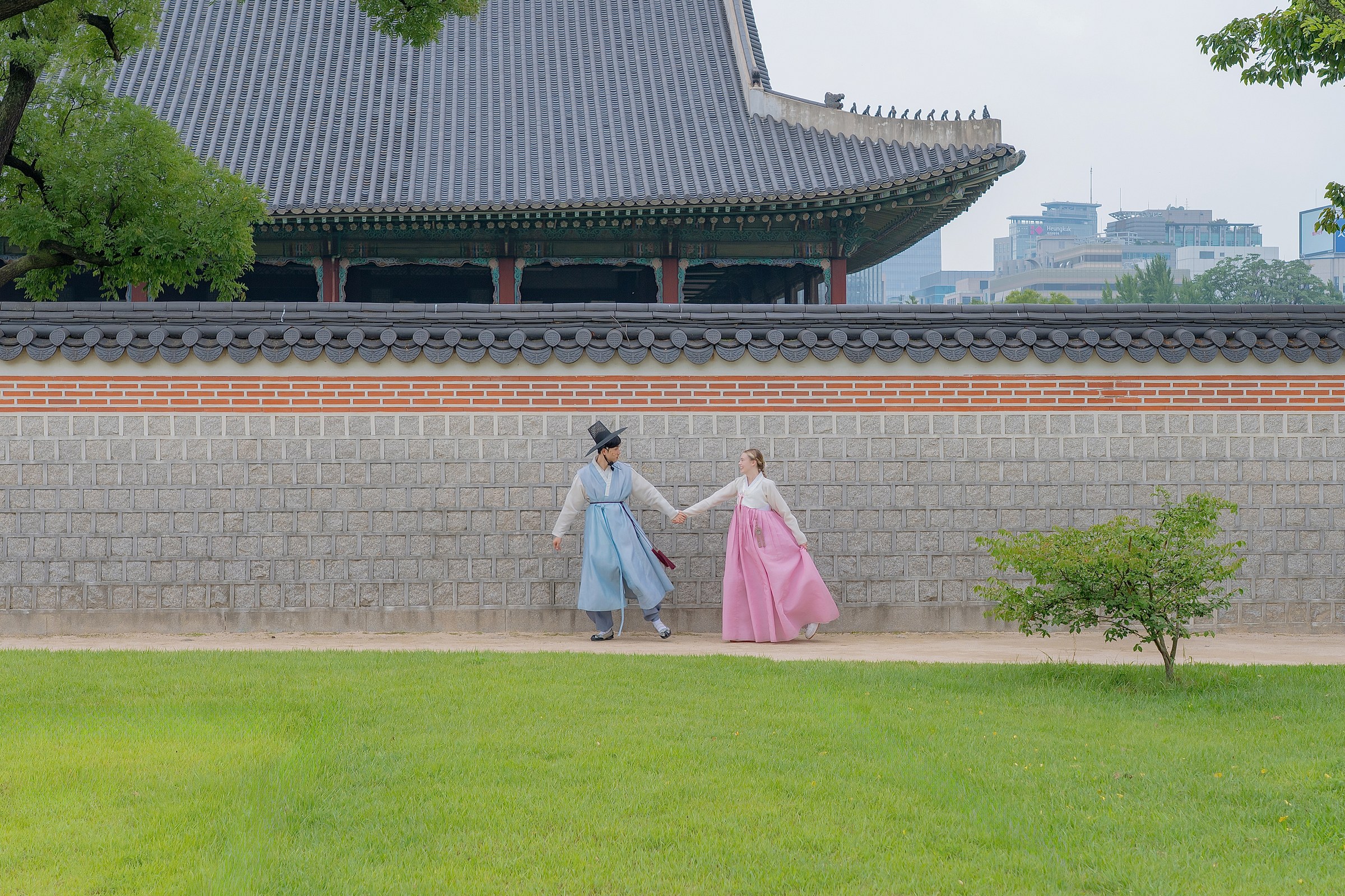 A couple in pastel hanbok walking hand in hand across a palace lawn