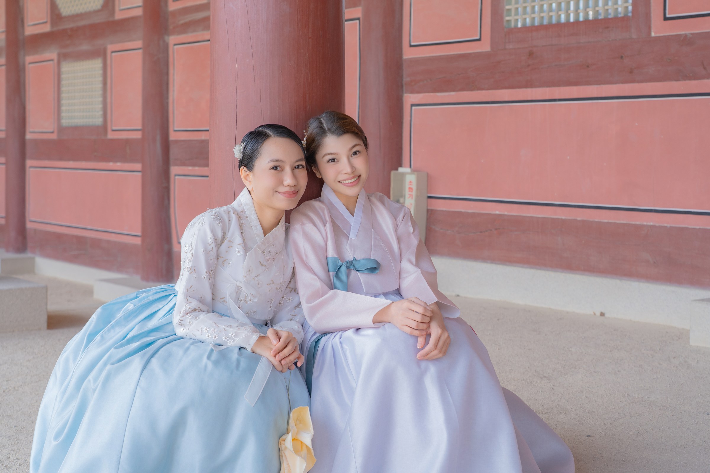 Two friends from the Philippines in blue and lavender hanbok seated beside a red palace wall